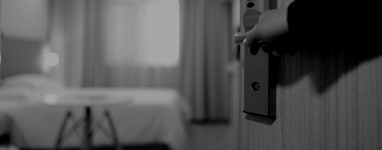 Black and white picture looking into a hotel room; a man's hand can be seen holding the door open